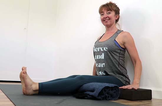 yoga teacher sitting in dandasana against a wall with legs supported and blocks under the hands.