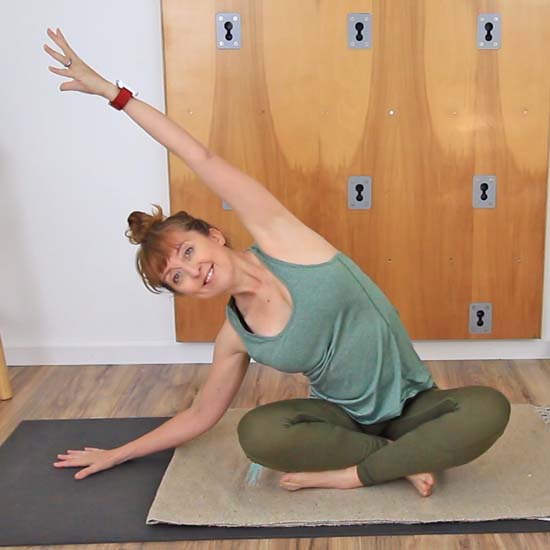 yoga teacher dressed in green seated on a  yoga mat reaching one arm up and over to the opposite side